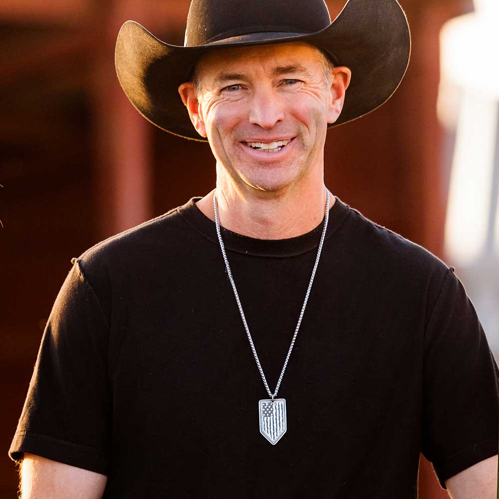 A man wears a gunmetal and silver arrow-shaped pendant featuring a tattered American flag design.