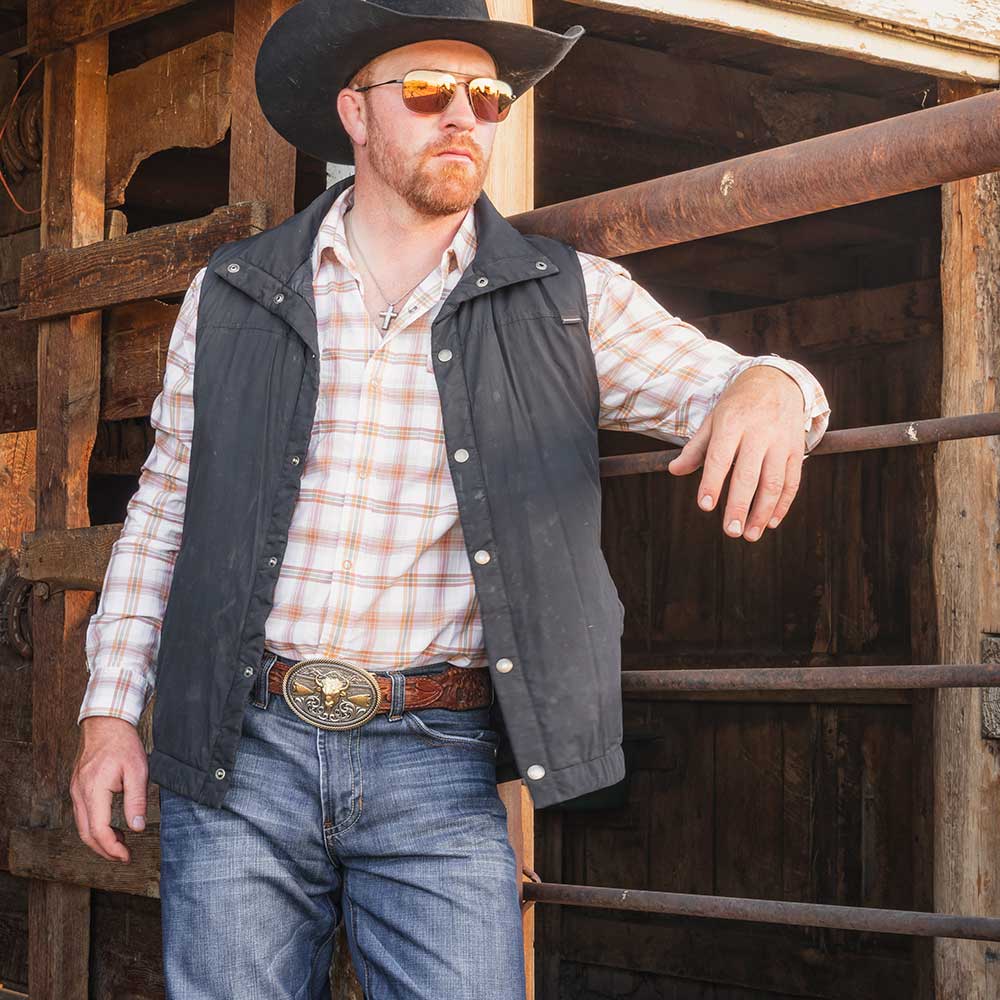 A man wearing a silver cross pendant necklace with a plaid shirt and black vest.