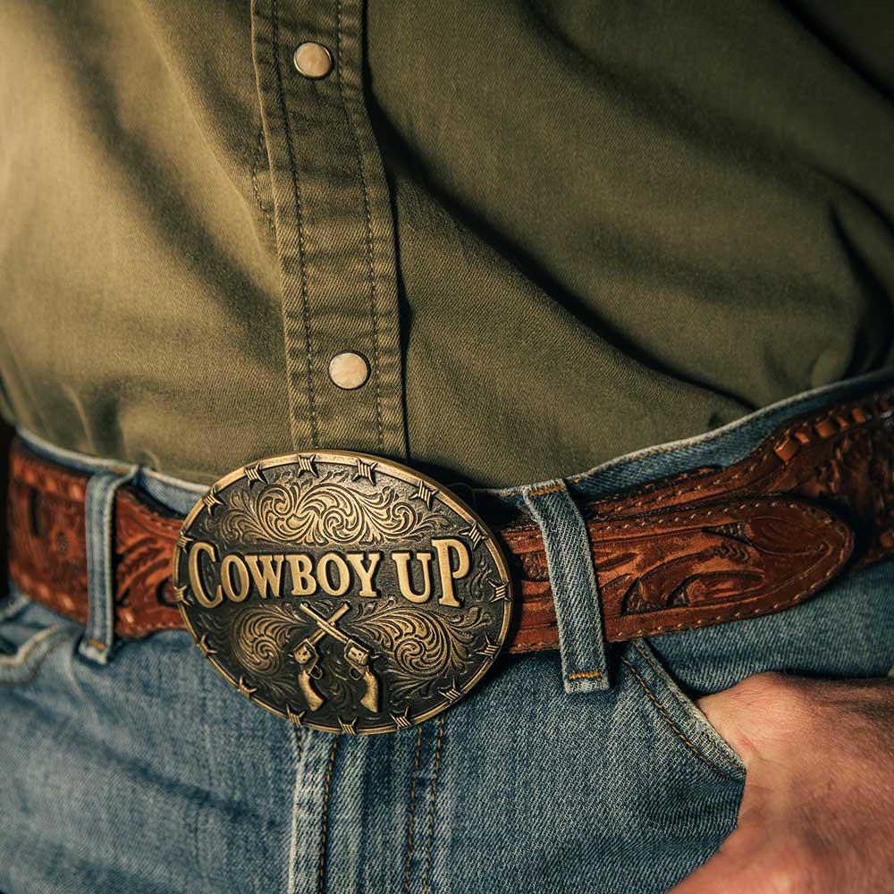 An oval gold-tone belt buckle featuring the words Cowboy Up and crossed revolvers.