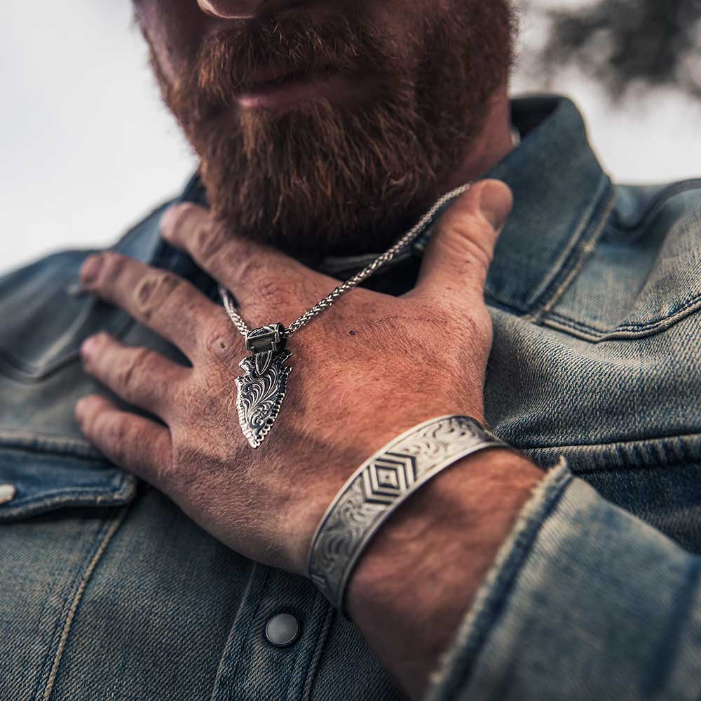 A man wears a gunmetal arrowhead pendant with Western engraving on a silver wheat chain.