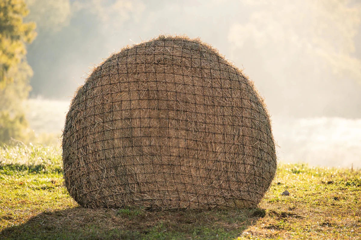 A round hay bale wrapped in a black UV-resistant nylon mesh net sitting in a grassy field.